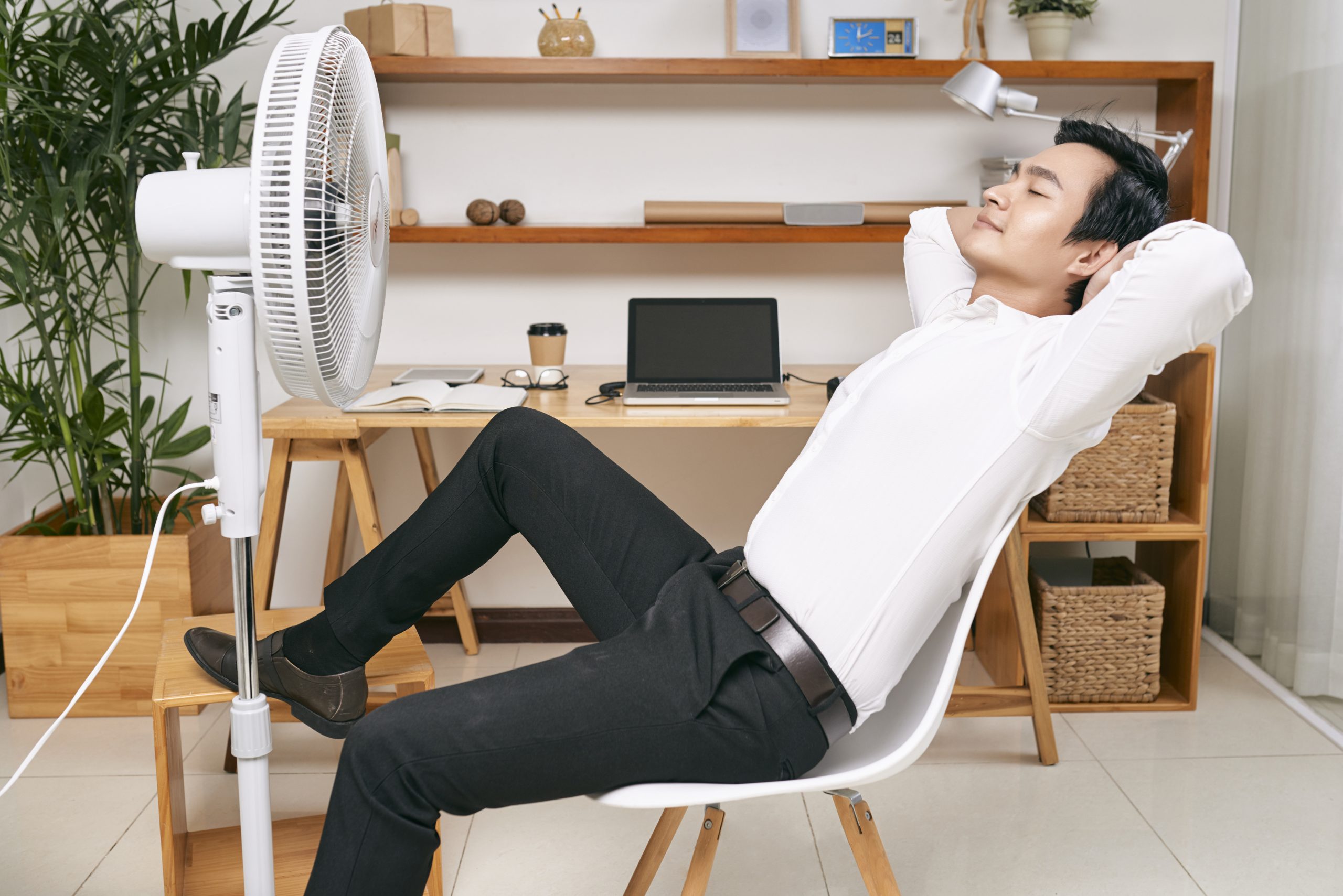 Young Asian office worker resting on chair in front of ventilator and refreshing from heat at office